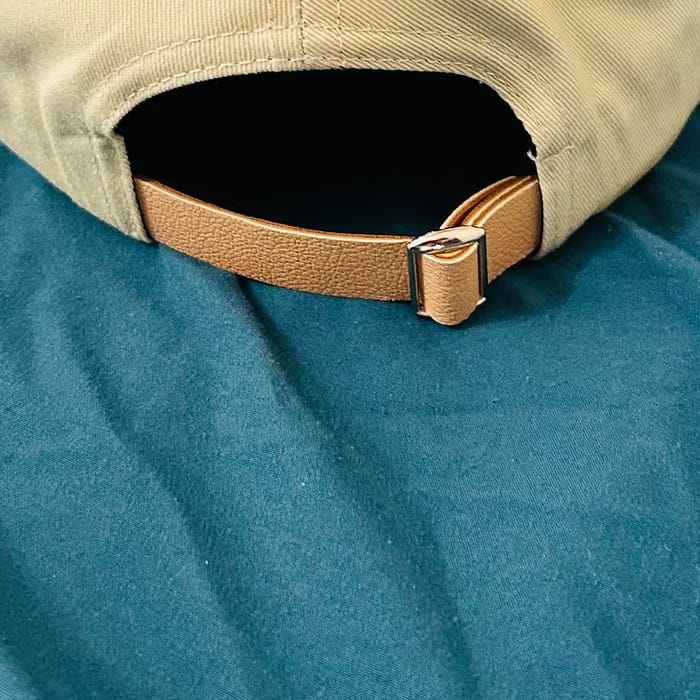 Tan leather adjustable strap with metal slider buckle on a cream baseball cap